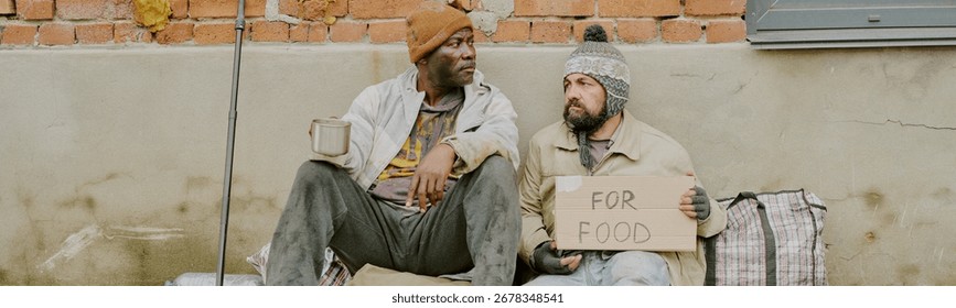 Two middle aged men, one Black and one Caucasian, sitting outdoors against brick wall, holding metal mug and cardboard sign reading for food, appearing to be homeless and seeking help