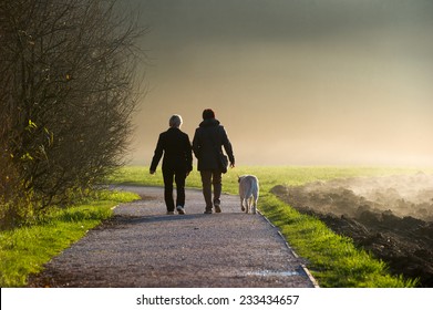 Two Middle Aged Women Walking A Dog On A Path In A Park.