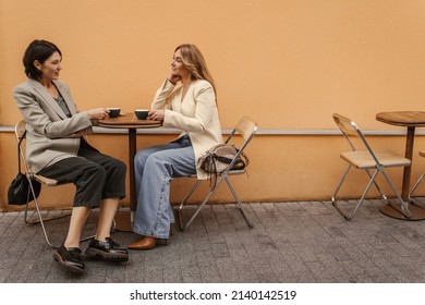 Two Middle-aged Caucasian Ladies Are Sitting At Table On Street, Chatting And Drinking Coffee, Tea. Brunette And Blonde In Casual Clothes Are Having Relaxed Time. Lifestyle And People Concept