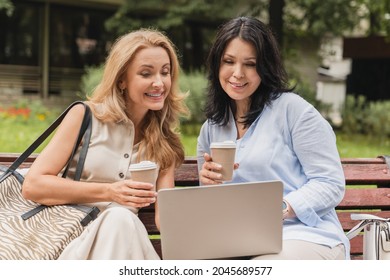 Two Middle-aged Mature Women Sisters Best Friends Girlfriends Watching Movie Webinar Working Online Remotely Together, Using E-banking E-commerce On Computer In City Park Drinking Coffee