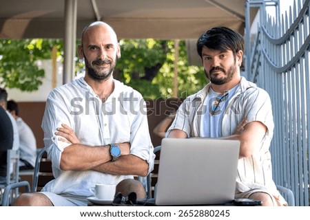 Two middle-aged men professionals happily work on their laptops in the garden of a cafe on a summer day