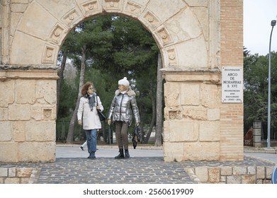 Two middle-aged women enjoying a leisurely walk under a historic stone archway in a picturesque Spanish village - Powered by Shutterstock