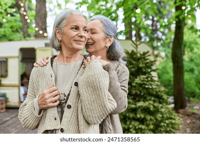 Two middle-aged women share a loving moment in a forest setting, near a camping van. - Powered by Shutterstock