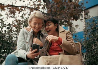 two middle-aged women are sitting outside using a mobile phone and are cheerful - Powered by Shutterstock