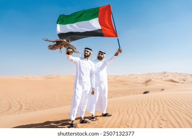 Two middle-eastern men wearing traditional emirati arab kandura bonding in the desert and holding a falcon bird and UAE flag - Arabian muslim friends meeting at the sand dunes in Dubai - Powered by Shutterstock