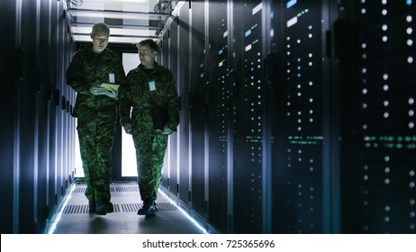 Two Military Men Walking In Data Center Corridor. One Uses Tablet Computer, They Have Discussion. Rows Of Working Data Servers By Their Sides.