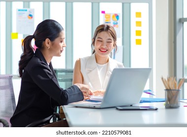 Two millennial Asian young beautiful professional successful businesswoman colleagues sitting smiling in meeting room having funny conversation together while working with laptop notebook computer.