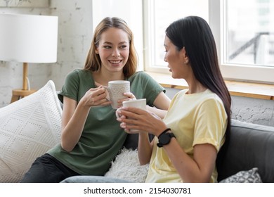 Two Millennial Girl Friends At Home On The Sofa Talking, Drinking Coffee, Discussing. Diverse Friends Spending Fun Time Together And Laughing, Chat And Talk. Asian And Caucasian Model.