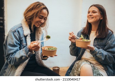 Two Millennial Women Female Friends Sitting Outdoors Eating Takeaway Food, Laughing And Having Fun. Food Delivery And Takeout.