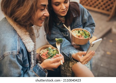 Two Millennial Women Female Friends Wearing Protective Face Mask Sitting Outdoors Eating Takeaway Food, Laughing And Having Fun. Food Delivery And Takeout.