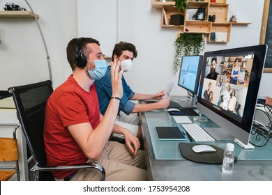 Two Millennials Men At Working In The Office Wearing Medical Mask For Protection By Coronavirus, Covid-19 Leader Greets In A Video Call Conference While The Colleague Near Him Working At The Computer