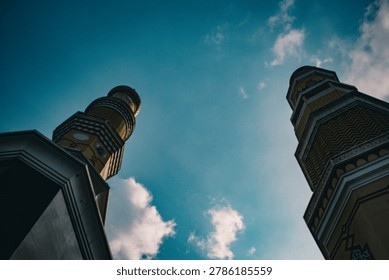 Two minarets of a mosque are viewed from below against a vibrant blue sky with scattered white clouds, creating a sense of grandeur.