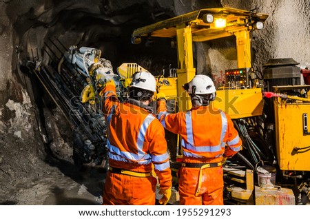 Two mine engineers with protective helmets standing and talking on a huge drill machine, talking and watching at the digging site. Rear view.