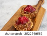 Two mini tarts with red fruit filling and a nut topping on a wooden cutting board.