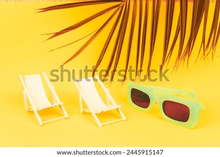Two miniature beach loungers on yellow background with sunglasses and palm leaf. Summer vacation concept.