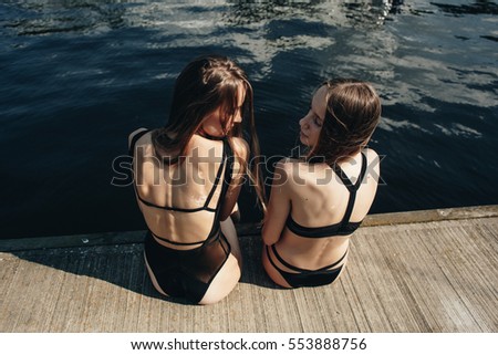 Two models in black swimsuit sitting back. Sea on background. Enjoying sunshine.