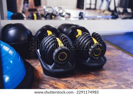Two modern adjustable dumbbells on stands. Sports equipment in the gym. The reflection of the mirror shows weighted ball, kettlebells and exercise equipment on background. Copy space.