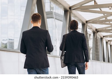 Two modern businesspeople view from the back walk across the city bridge against the background of urban offices and buildings