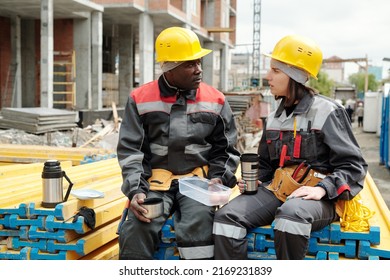 Two Modern Engineers Or Builders With Cups Of Hot Tea Having Talk At Construction Site While Sitting On Stack Of Yellow Building Materials