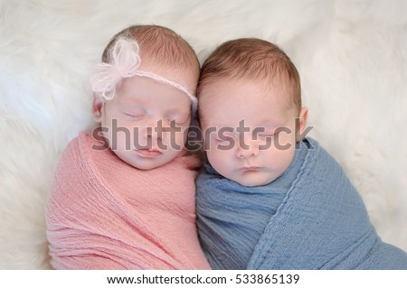 Two month old, fraternal twin, brother and sister babies swaddled in pink and blue wraps and sleeping on a sheepskin rug.