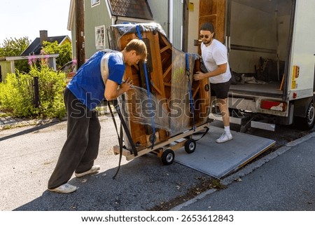 Two movers loading a partially wrapped grand piano onto a truck using a hydraulic lift on a residential street.