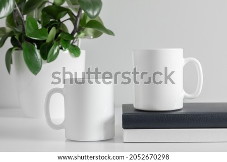 Two mugs mockup with notebook and green plant in pot on white table. front view
