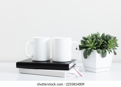 Two mugs mockup with notepad, accessories on white table and green plant in pot. Front view. - Powered by Shutterstock