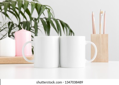 Two mugs mockup with workspace accessories and candles on a white table and a palm plant.