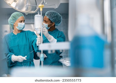 Two multi ethnic female surgeons having discussion and preparing Intravenous therapy for operation while wearing operating gowns, face masks and surgical gloves in emergency room in hospital. - Powered by Shutterstock