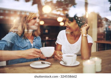 Two Multi Ethnic Friends Enjoying Coffee Together In A Coffee Shop Viewed Through Glass With Reflections As They Sit At A Table Chatting And Laughing