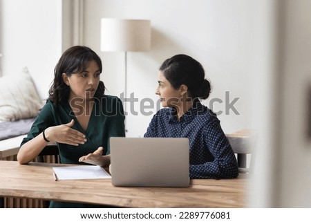 Two multi-ethnic businesswomen colleagues sit at desk, discuss joint project sit in office at workplace desk with laptop, share vision, ideas, planning cooperation. Teamwork, apprenticeship, mentoring