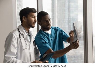 Two multiethnic doctors radiologists or surgeons dressed in white coat in blue scrubs examining X-ray, discussing patient condition, diagnoses, or treatment options based on images findings. Teamwork - Powered by Shutterstock