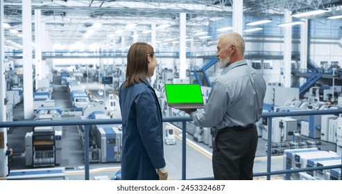 Two Multiethnic Engineers Standing on a Platform, Using Laptop Computer with Green Screen at an Electronics Factory. Machines are Undergoing Maintenance, Specialists Monitoring the Progress - Powered by Shutterstock