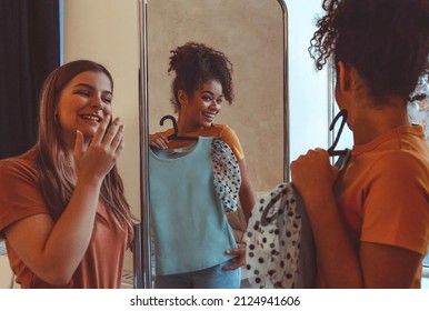 Two Multiethnic Girlfriends Having Fun Trying On Clothes In Front Of Mirror At Home, Smiling African American Girl Holds Dress On Hanger Showing Outfit To Female Friend Standing Next. Friendship