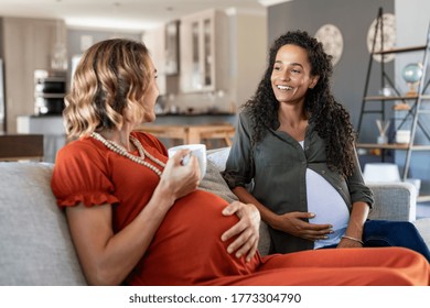 Two multiethnic pregnant women enjoying tea time at home. Smiling pregnant women talking and looking at each other. Happy girls both with a baby bump relaxing on couch and have a friendly conversation - Powered by Shutterstock