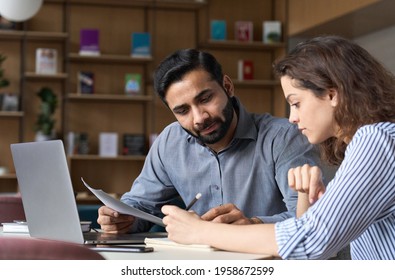 Two Multiethnic Professionals Colleagues Working Together With Laptop And Papers In Office. Indian Male Mentor And Latin Female Young Professional Sitting In Creative Office Space.