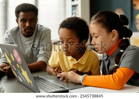 Two multiethnic younger students using laptop together keeping fingers on touchpad fully engaged in educational game on screen, while male teacher helping kids sitting at desk