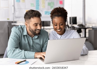 Two multinational smiling colleagues looking at laptop screen seated together at desk in modern office, collaborating on project, reviewing data, or watching amusing content on computer during break