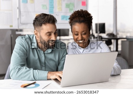 Two multinational smiling colleagues looking at laptop screen seated together at desk in modern office, collaborating on project, reviewing data, or watching amusing content on computer during break