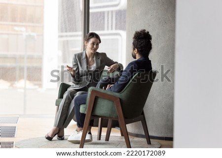 Two multinational workmates talking seated on armchairs in modern office, share opinion and thoughts, discuss common project, share solution looking interested in conversation. Business, communication