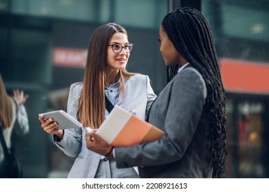 Two Multiracial Business Woman Walking Outside Of Office Buildings. Young Diverse Females Professional In The City. Woman Leader In A Business. Focus On A Caucasian Woman Holding Digital Tablet.