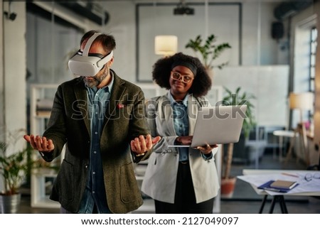 Two multiracial coworkers brainstorming together in the office. Man wearing VR headsets while his female african colleague standing beside and holding laptop
