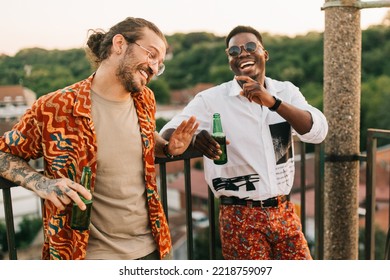 Two multiracial friends have fun at the open-air summer rooftop party, tell jokes and laugh while holding a beer and leaning on the railing.