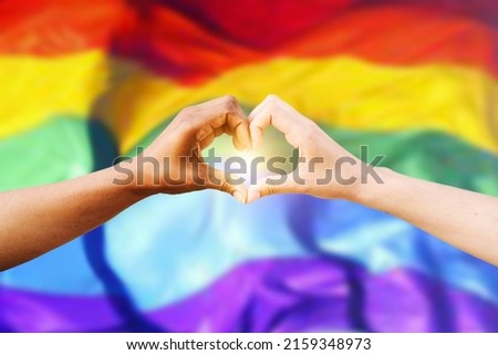 Two multiracial hands join together to form a shape heart with a LGBT flag background. Concept of homosexual gay pride day and love between different races, peace, humans against discrimination