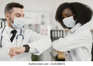Two Multiracial Male And Female Doctors In Face Masks Doing Elbow Bump During Meeting A Hospital Room. New Normal Of Greeting During Coronavirus.