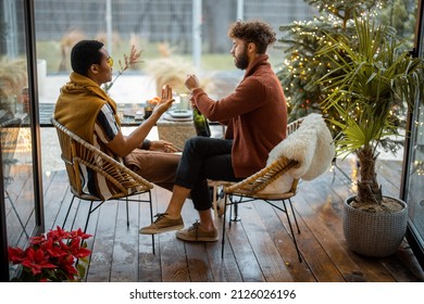 Two Multiracial Male Friends Drinking Spakling Wine, Celebrating Winter Holidays, Sitting By A Dinner Table At Backyard With Christmass Tree. Caucasian And Hispanic Man Having A Festive Dinner