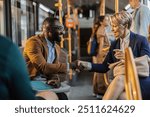 Two multiracial, middle-aged business professionals shake hands and smile while sitting on a public bus, signifying a positive interaction.