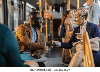 Two multiracial, middle-aged business professionals shake hands and smile while sitting on a public bus, signifying a positive interaction. - Powered by Shutterstock