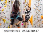 Two multiracial women doing rock climbing in the gym. Caucasian and asian girls climb the bouldering wall. Professional sport female climber train. Cute multiethnic friends are actively spending time.