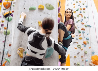 Two multiracial women doing rock climbing in the gym. Caucasian and asian girls climb the bouldering wall. Professional sport female climber train. Cute multiethnic friends are actively spending time. - Powered by Shutterstock